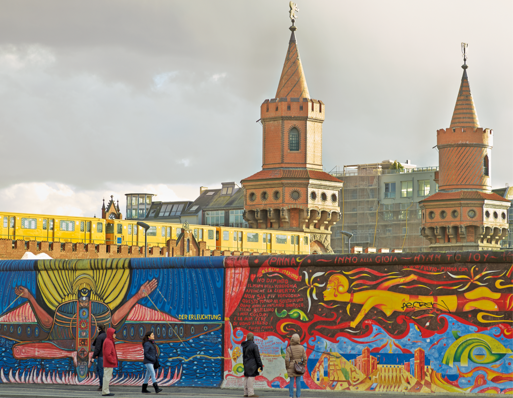 Fotografia. Pessoas observando desenhos e grafites coloridos em uma parte do Muro de Berlim. Ao fundo, atrás do muro, duas torres e prédios.