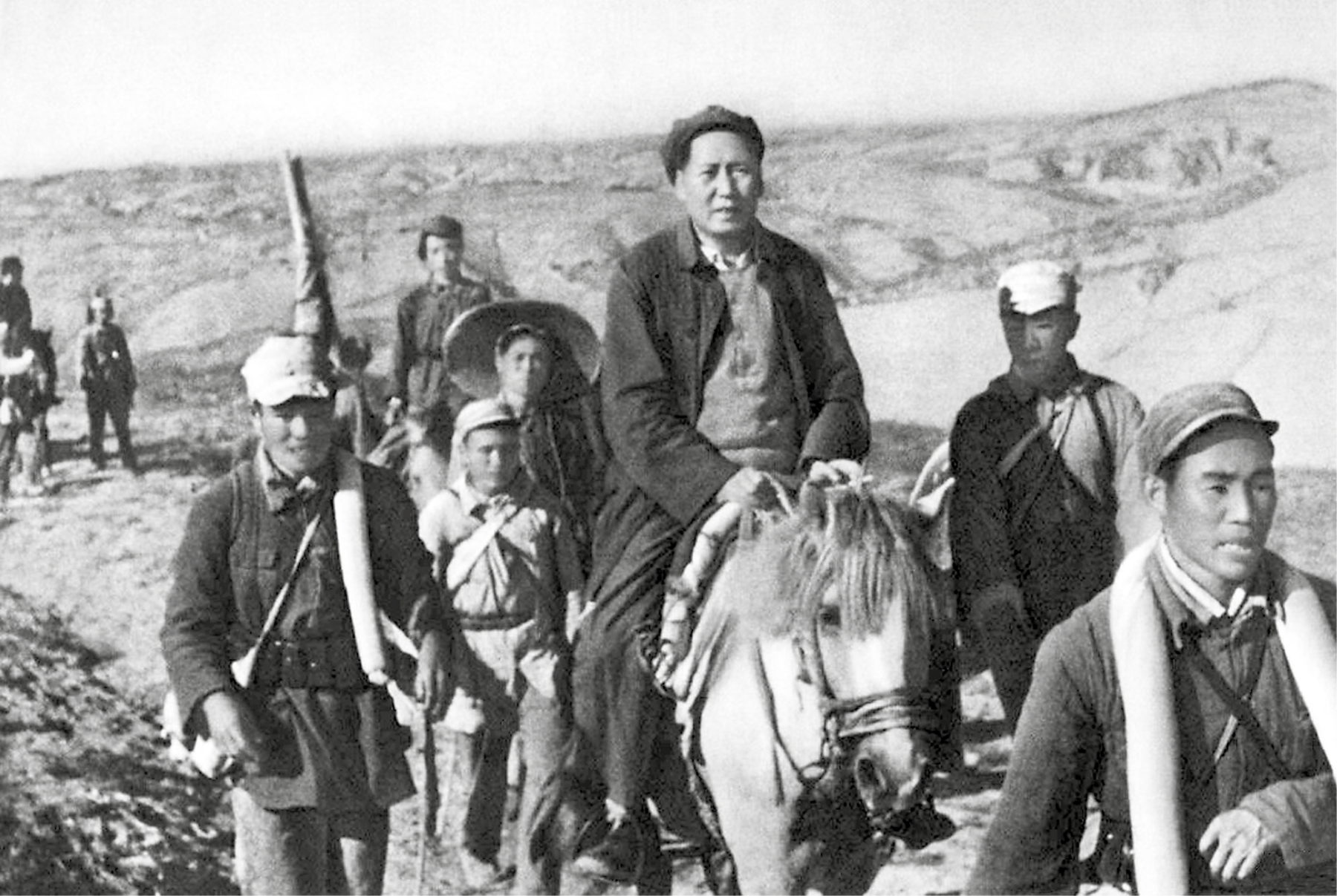 Fotografia em preto e branco. Mao Tsé-tung, homem de casaco e boina montado em um cavalo. Ao redor dele, pessoas caminham.