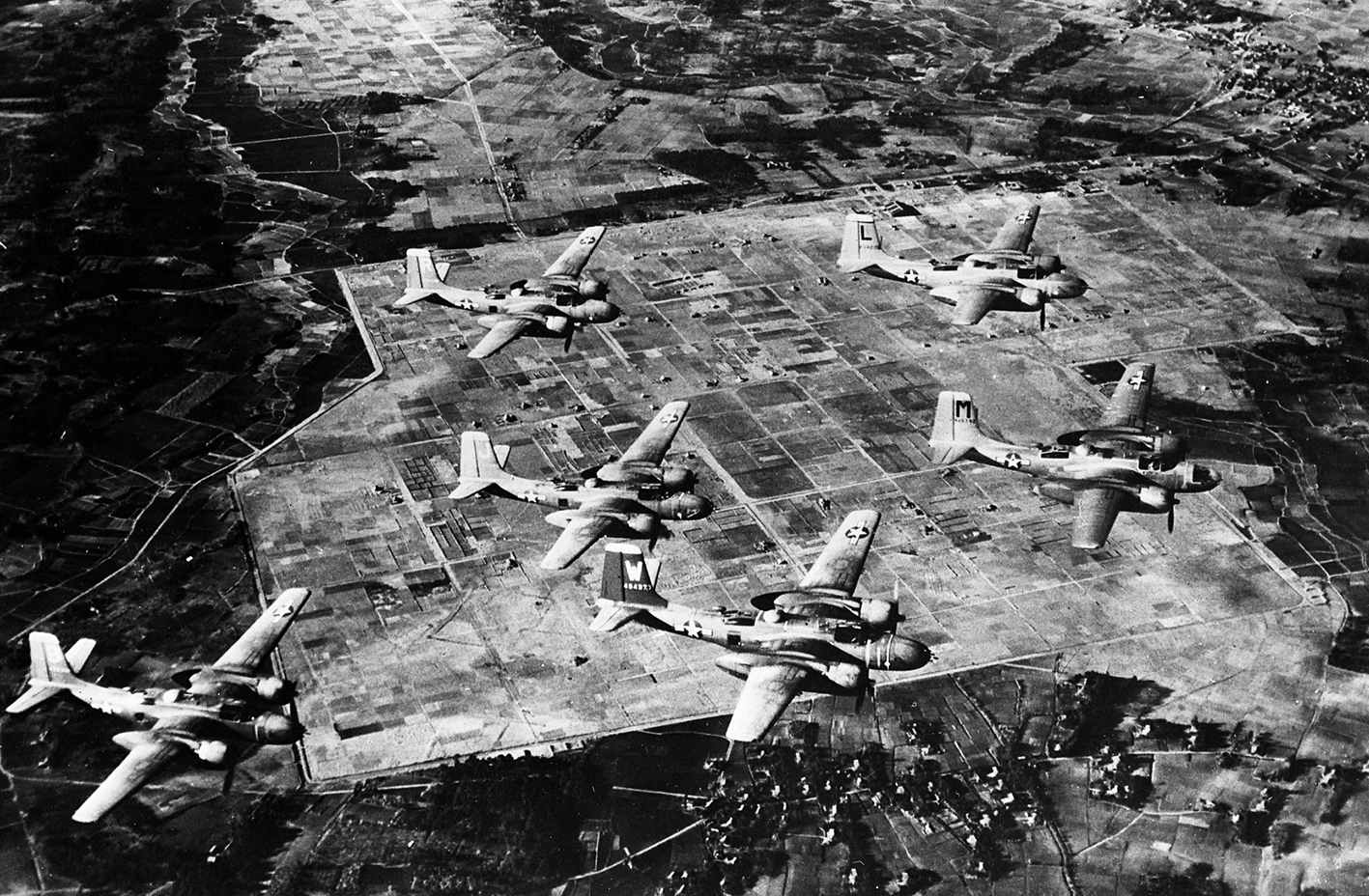 Fotografia aérea em preto e branco. Aviões sobrevoam plantações e vilarejos.