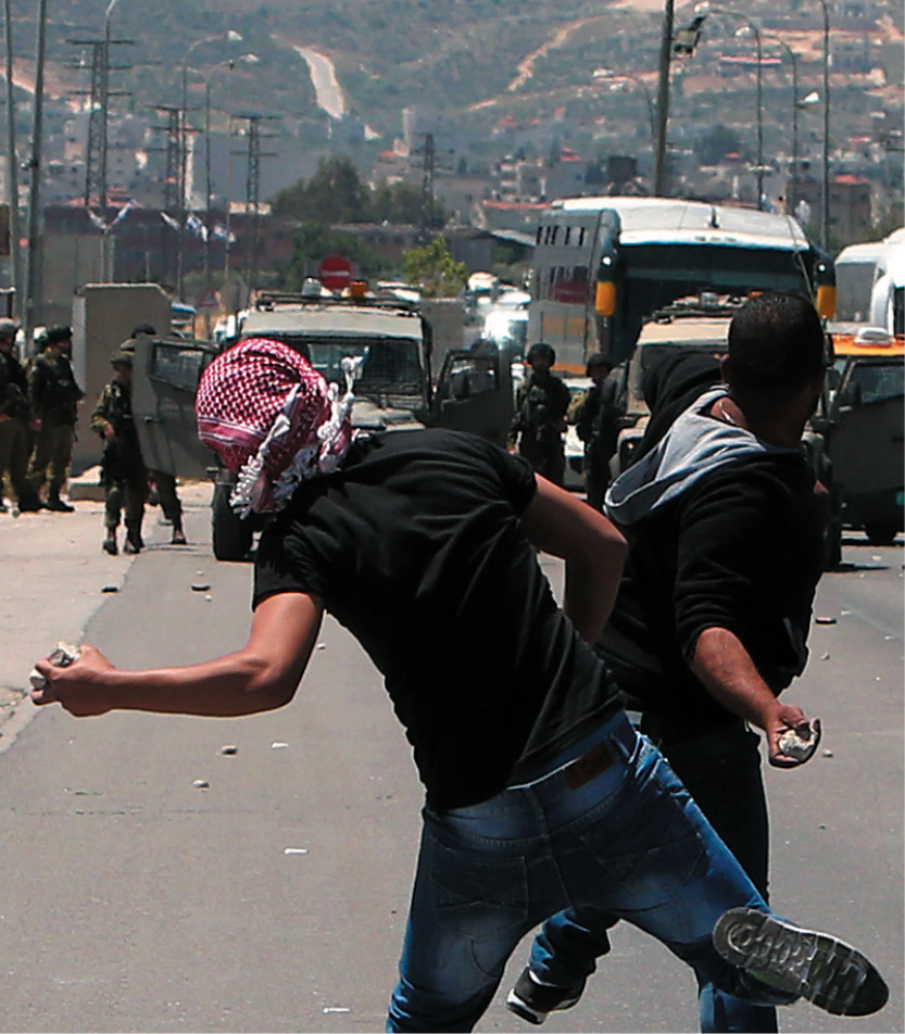 Fotografia. Manifestantes atirando pedras em militares que estão do outro lado da rua.