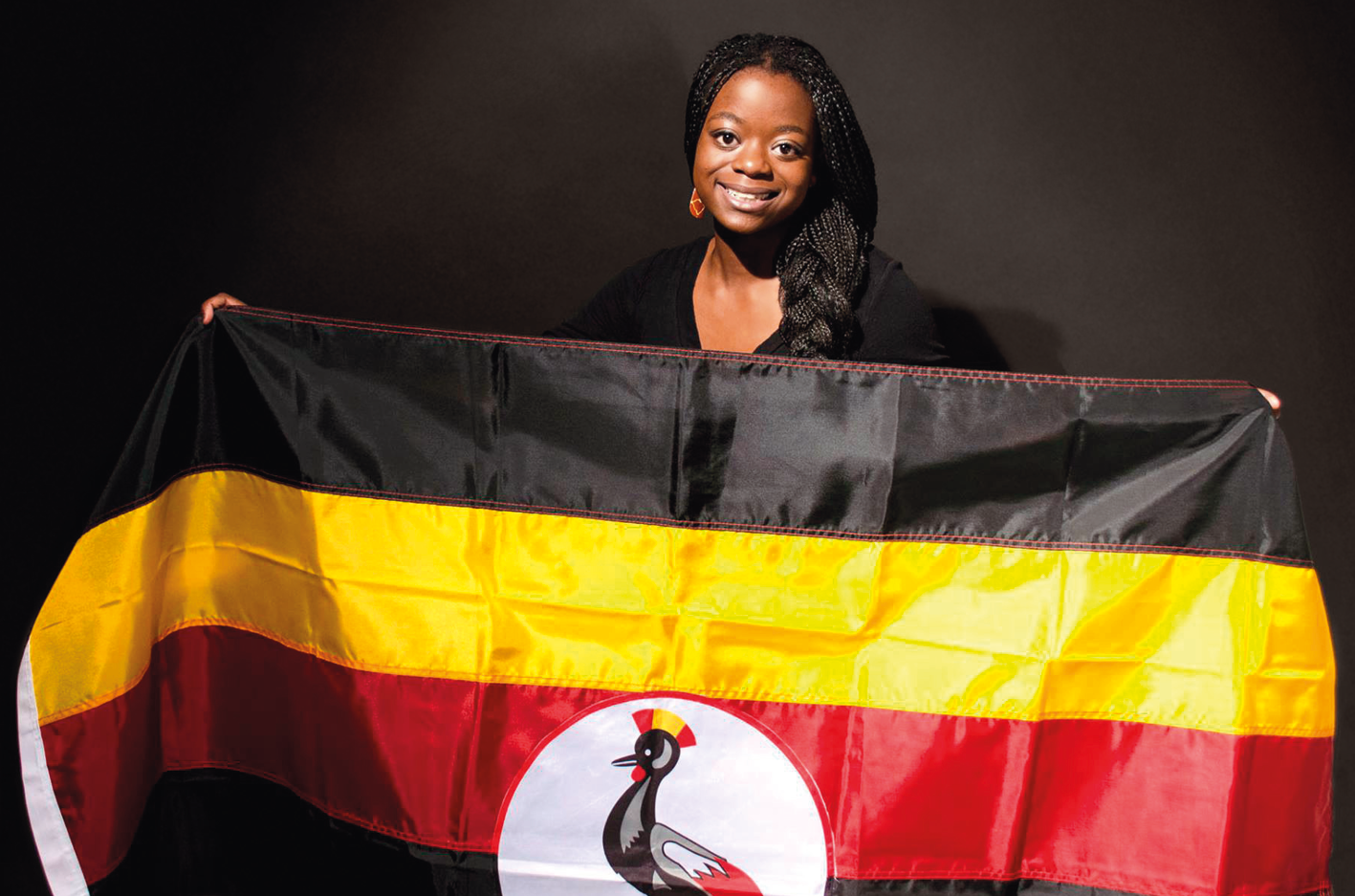 Fotografia. Mulher sorridente, segurando a bandeira de Uganda. Composta por faixa horizontais, em preto, amarelo, vermelho e preto. No centro um círculo branco com uma ave preta desenhada.