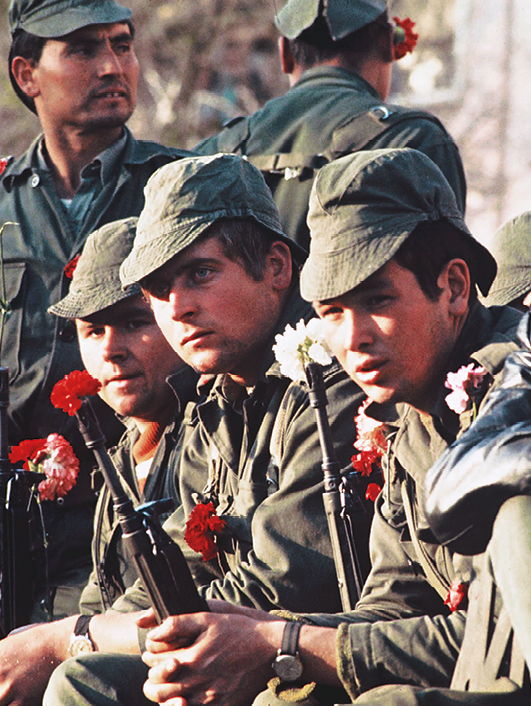 Fotografia. Soldados sentados segurando armas compridas. Dentro dos canos das armas, cravos vermelhos e brancos.