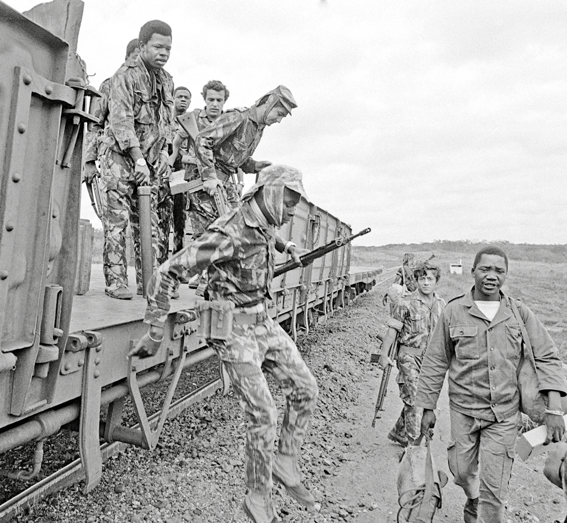 Fotografia em preto e branco. Militares descendo de um trem. Alguns caminham ao lado do trilho, em uma região árida. Alguns carregam armas.