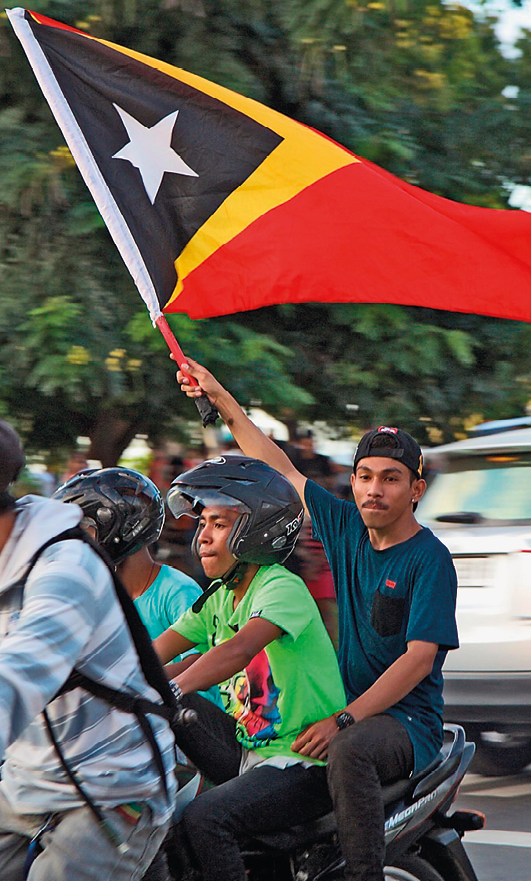 Fotografia. Dois rapazes em uma moto. O garupa está sem capacete e carrega uma bandeira do Timor Leste.