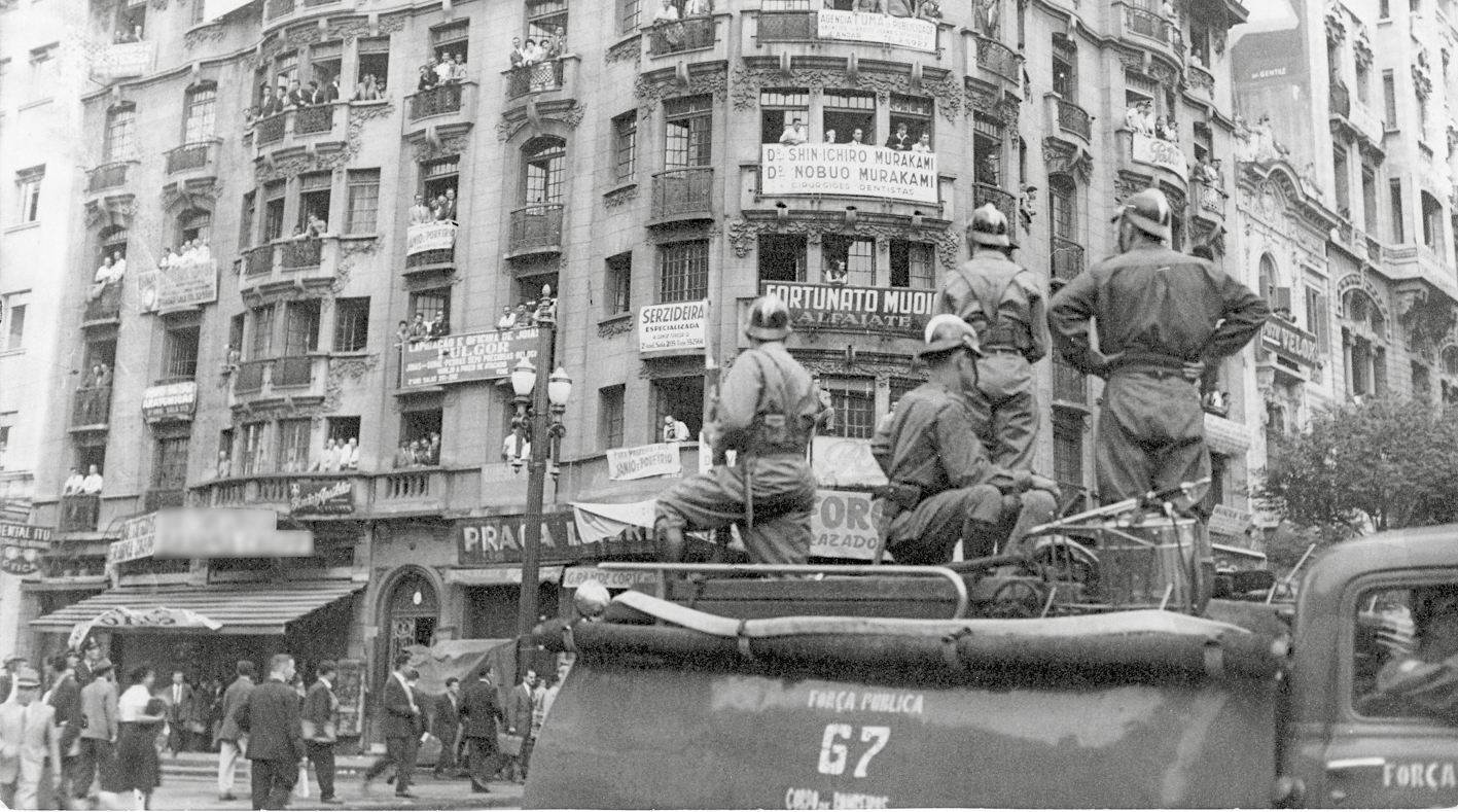 Fotografia em preto e branco. Militares dentro de um carro em uma rua movimentada. Ao fundo, prédio grande, de esquina com muitas pessoas nas janelas.
