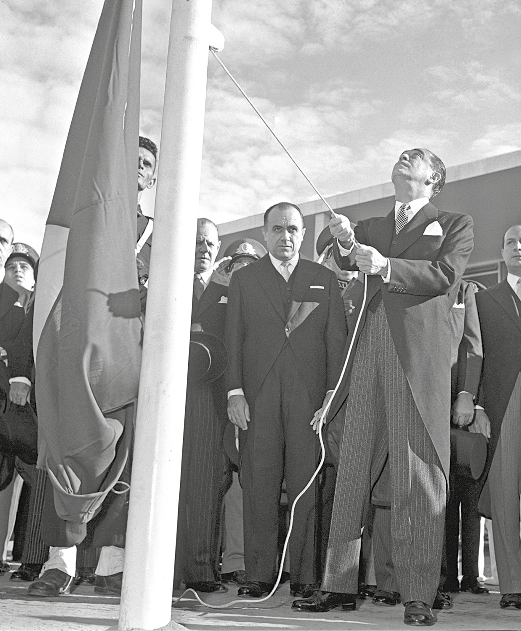 Fotografia em preto e branco. Juscelino Kubitschek hasteando uma bandeira. Ele está olhando para cima. Ao redor dele pessoas observam o ato.