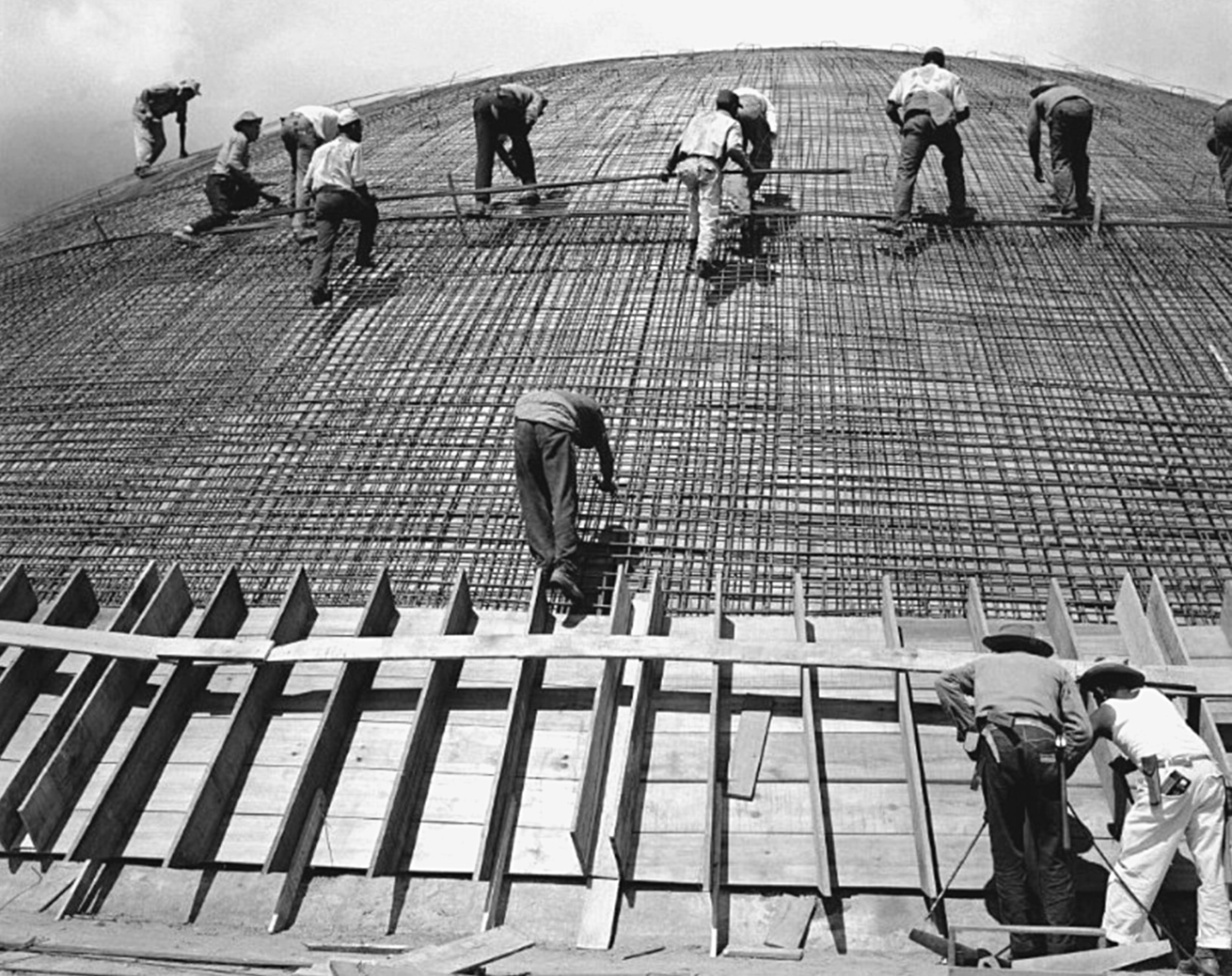 Fotografia em preto e branco. Operários trabalhando em uma grande construção com diversas vigas de ferro entrelaçadas.