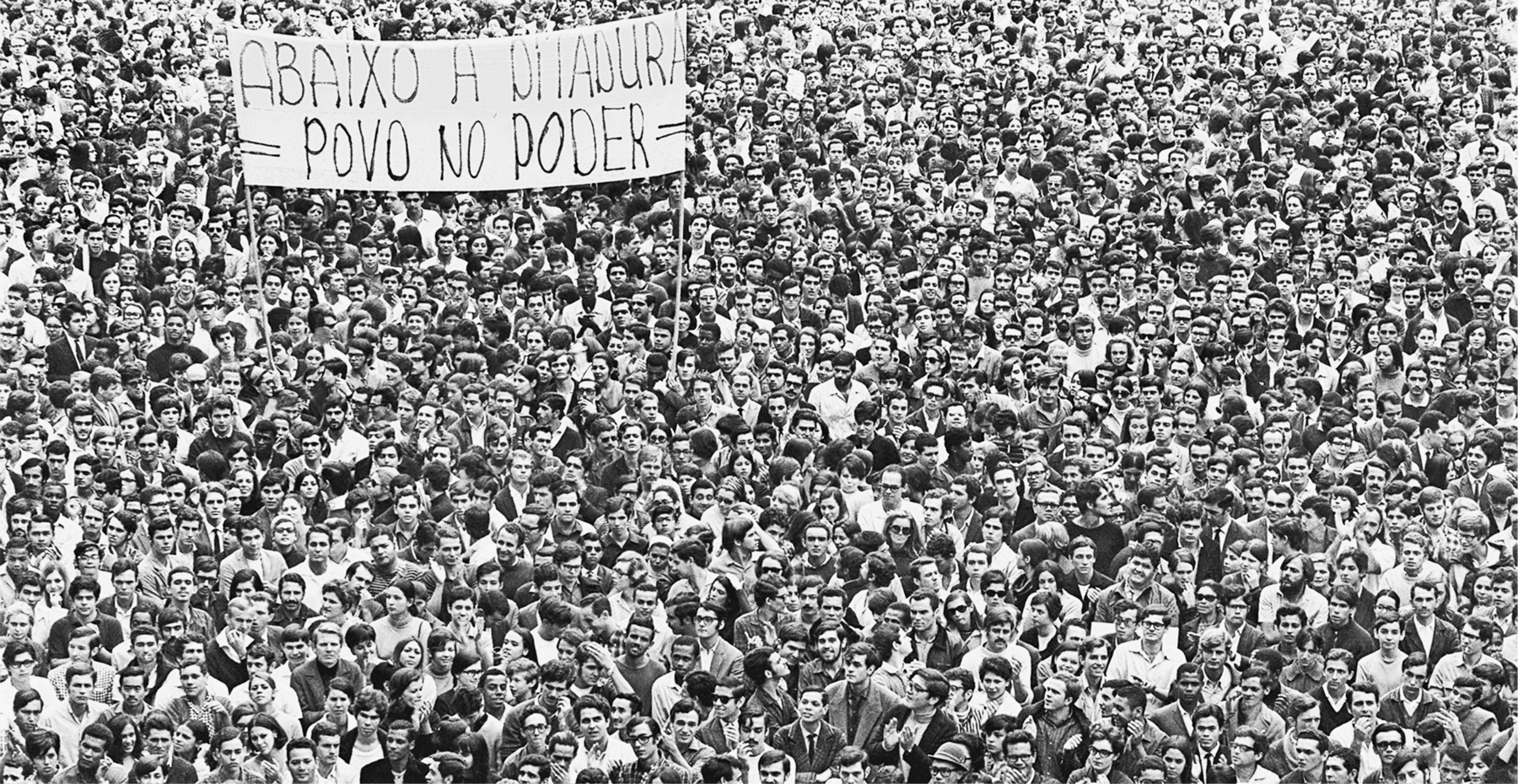 Fotografia em preto e branco. Multidão em uma passeata, carregando um cartaz com a frase: ABAIXO A DITADURA – POVO NO PODER.