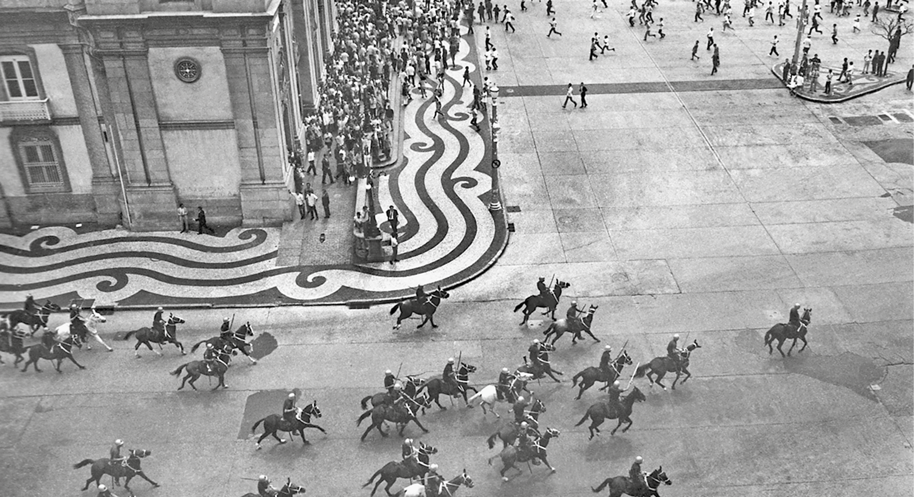 Fotografia em preto e branco. Militares montados em cavalos dispersam a multidão em uma larga avenida.