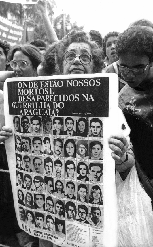 Fotografia em preto e branco. Alzira Grabois, senhora de cabelos curtos e óculos, carrega um cartaz com retratos de pessoas desaparecidas. No cartaz, acima das fotos, está escrito: ONDE ESTÃO NOSSOS MORTOS E DESAPARECIDOS NA GUERRILHA DO ARAGUAIA?