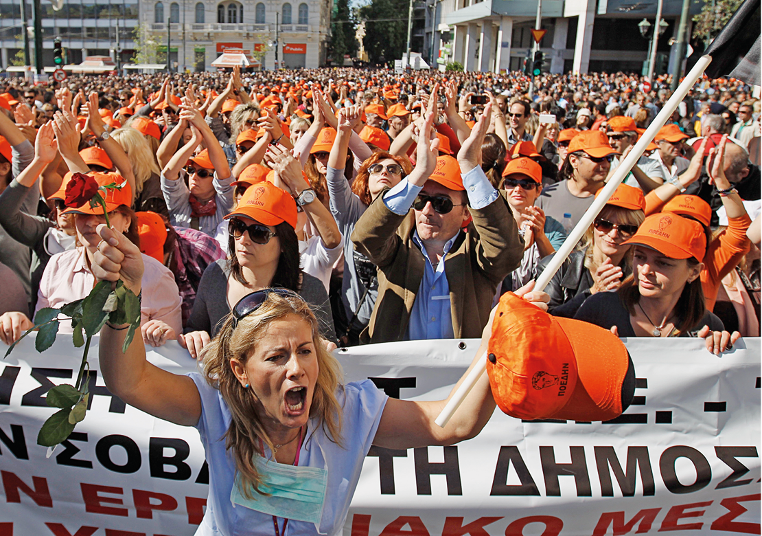 Fotografia. Multidão reunida em uma manifestação de rua. Todos usam boné laranja. À frente, uma longa faixa e uma mulher com a boca aberta, segurando o boné na mão.
