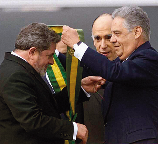 Fotografia. Fernando Henrique colocando a faixa presidencial em Lula. Estão um de frente para o outro. Lula está com a cabeça abaixada passando o braço esquerdo por dentro da faixa.