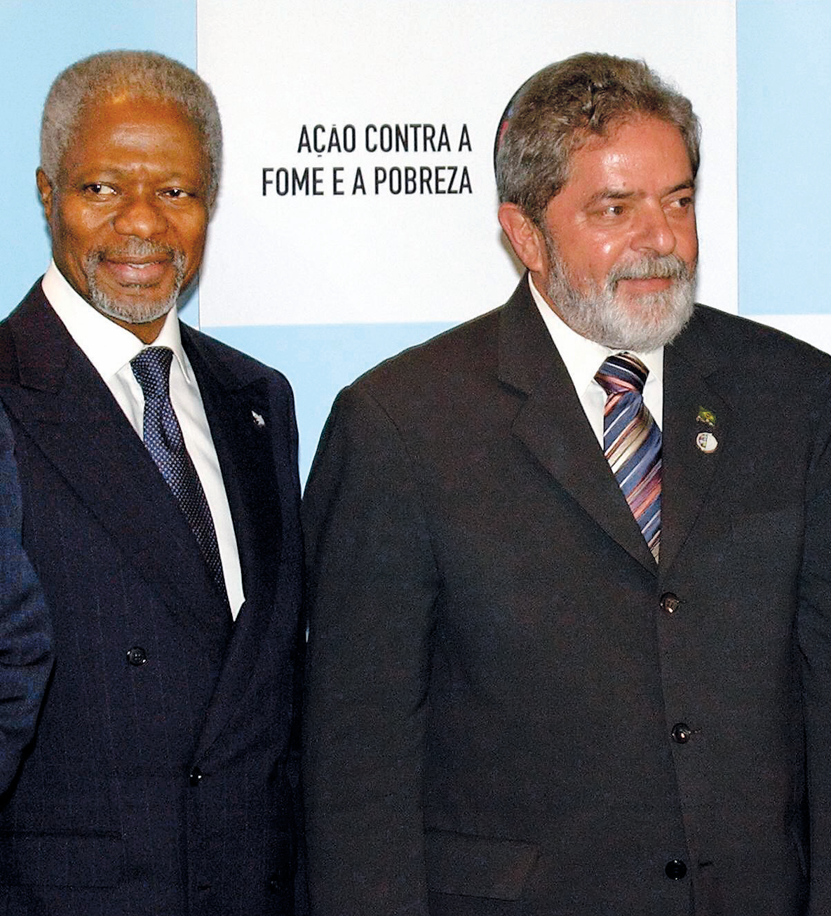 Fotografia. Kofi Annan, homem de cabelo e barba grisalhos. Veste terno azul escuro. Está ao lado de Lula, homem de cabelo e barba grisalhos. Veste terno preto e gravata colorida listrada. Atrás deles, na parede, está escrito: AÇÃO CONTRA A FOME E A POBREZA.