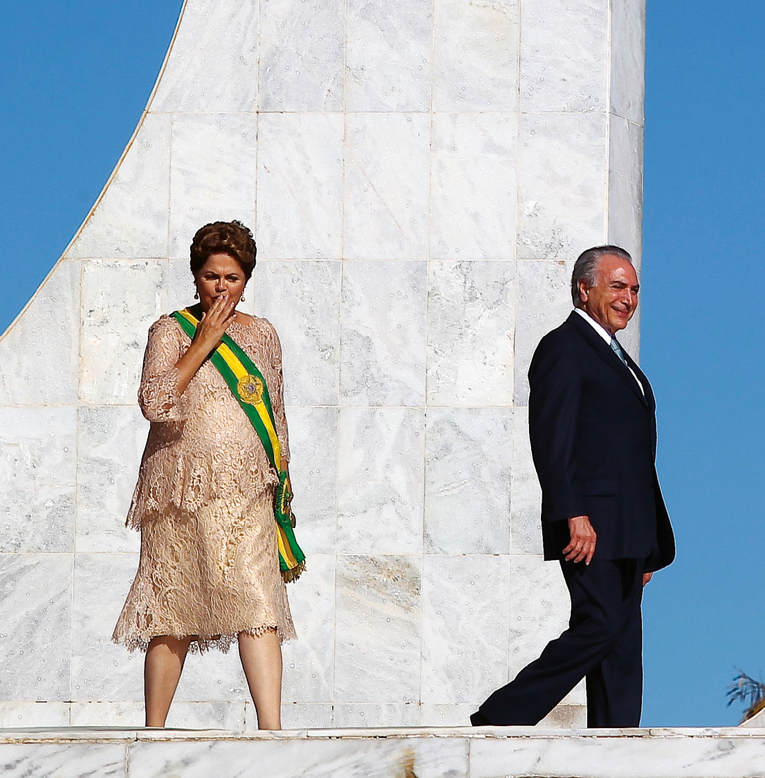 Fotografia. Dilma Rousseff, mulher de cabelos curtos e castanhos, usando um vestido largo e a faixa presidencial. Está com uma das mãos à frente da boca. Ao seu lado, de perfil, Michel temer, homem de cabelos grisalhos. Usa terno escuro.