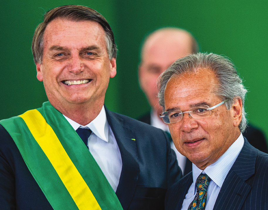 Fotografia. Jair Bolsonaro, homem sorridente, de cabeça pequena, cabelo liso penteado de lado. Tem os olhos pequenos. Está usando a faixa presidencial, com uma listra amarela entre duas verdes. Ao lado dele, Paulo Guedes, homem baixo, calvo e grisalho. Tem o rosto arredondado e usa óculos.