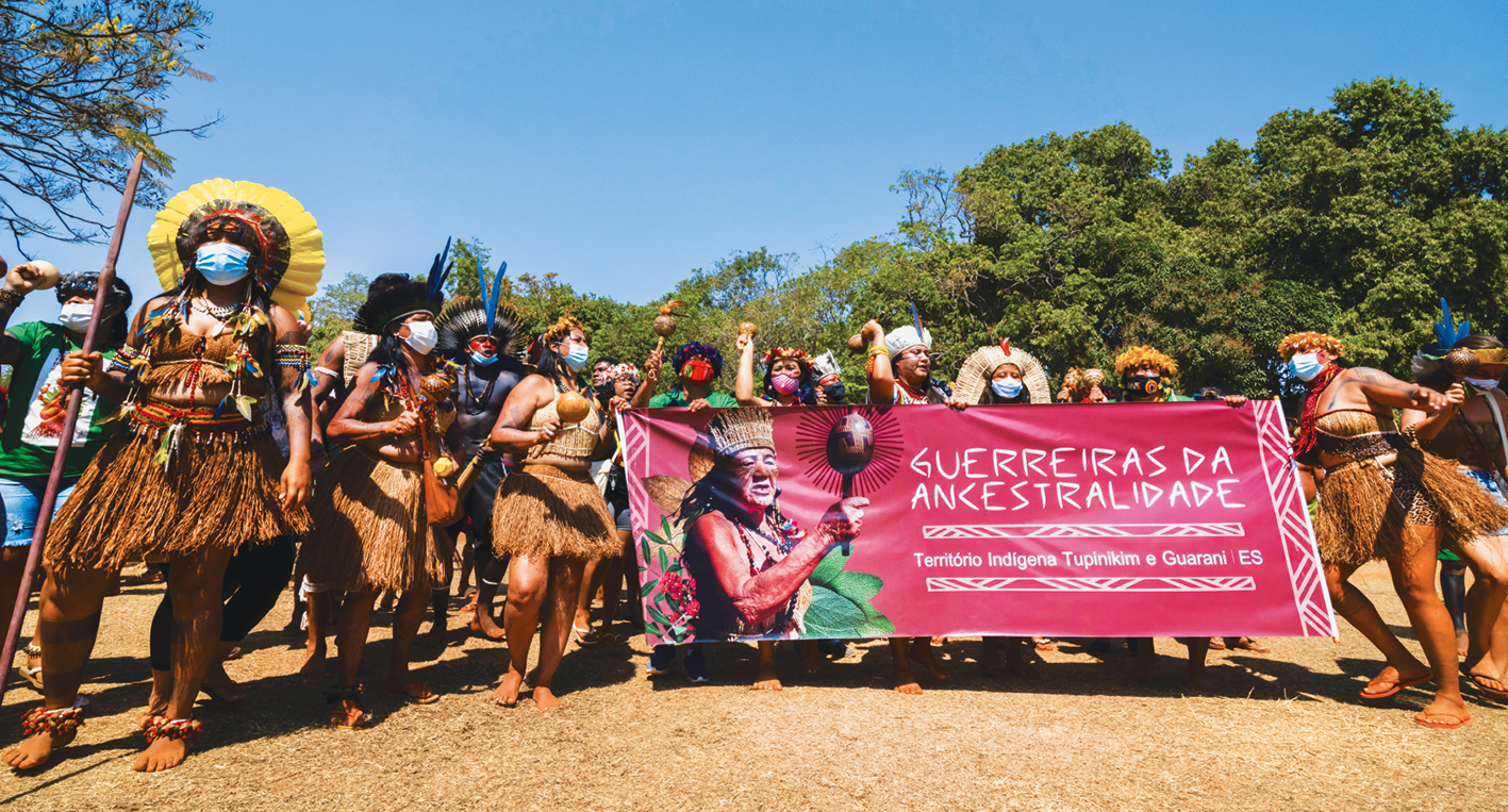 Fotografia. Mulheres indígenas em uma manifestação. Elas usam máscara de proteção facial. Carregam um cartaz com os dizeres: GUERREIRAS DA ANCESTRALIDADE. Ao fundo, se vê copas de árvores.