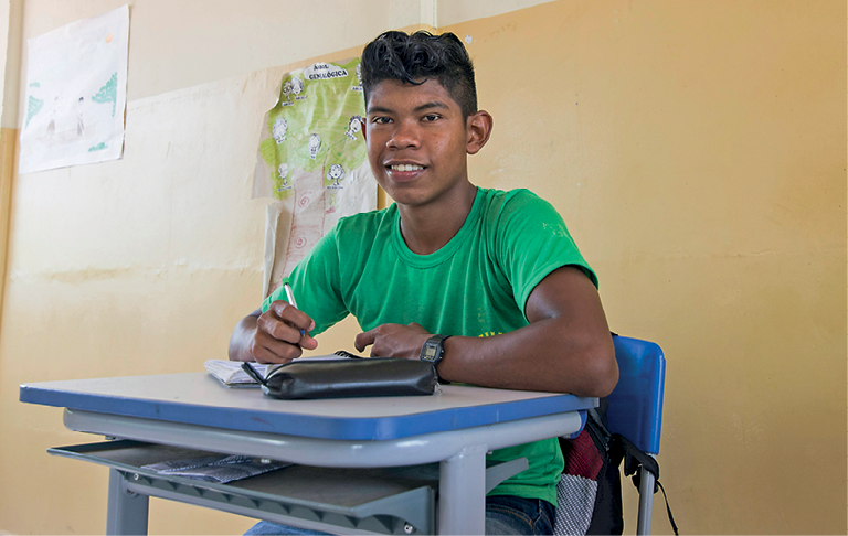 Fotografia. Um garoto sentado em cadeira de frente para carteira escolar em cinza e azul. Ele tem cabelos pretos, com camiseta verde, olhando para frente, sorrindo, com caderno e livro sobre a carteira, escrevendo com a mão esquerda. Ele olha para frente. Ao fundo, há uma parede de cor bege.