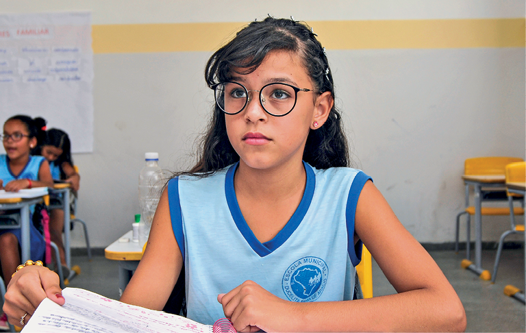 Fotografia. Uma menina sentada de frente para carteira escolar, vista da cintura para cima. Ela tem cabelos longos escuros com franja para a esquerda, usando óculos de grau redondos, de regata azul com contornos em azul mais escuro na gola e nas mangas. Ela segura na mão esquerda uma folha de caderno e olha para frente. Ao fundo, à esquerda, vista parcial de carteiras com outras crianças sentadas.