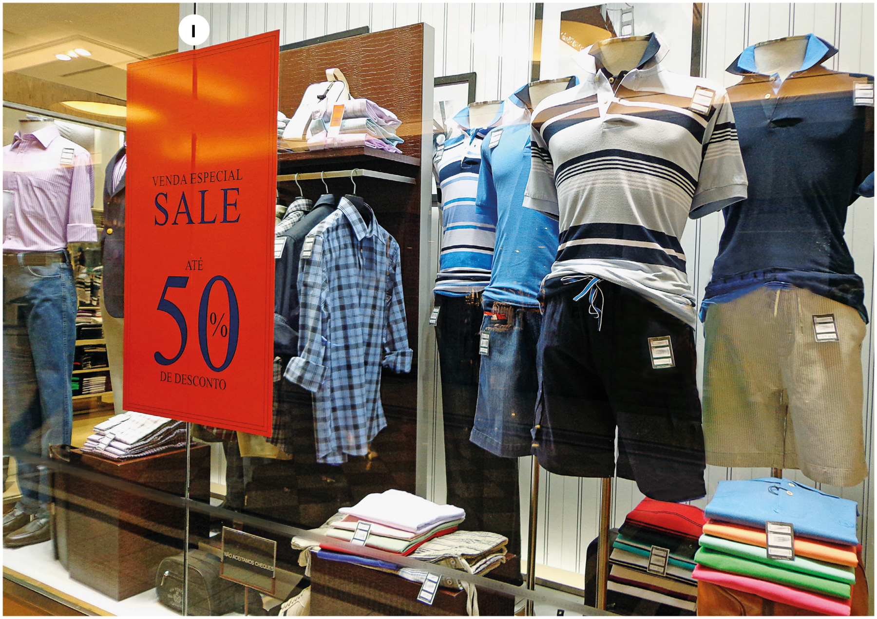 Fotografia um. Vista geral de uma vitrine. Nela há manequins usando camiseta, bermuda e mais ao fundo, camiseta de e calça jeans. Na parte inferior há camisetas dobradas uma sobre a outra, formando pilhas. Sobre a vitrine, adesivo em vermelho, escrito em azul: VENDA ESPECIA,L SALE, ATÉ CINQUENTA POR CENTO DE DESCONTO.