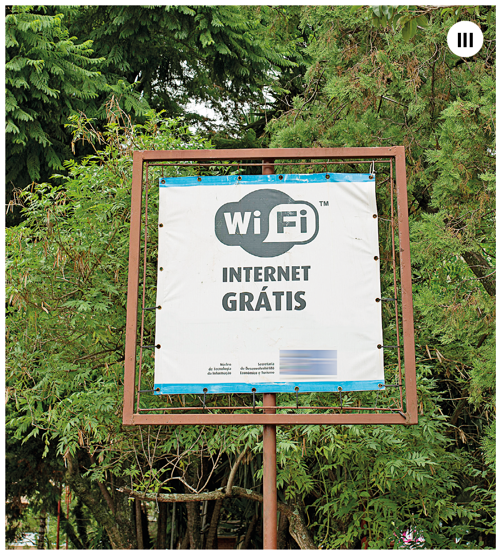 Fotografia três. Uma haste na vertical em marrom, e no topo, um cartaz em cor branca e com texto em preto: WIFI Internet Grátis. Ao fundo, árvores de folhas em verde.