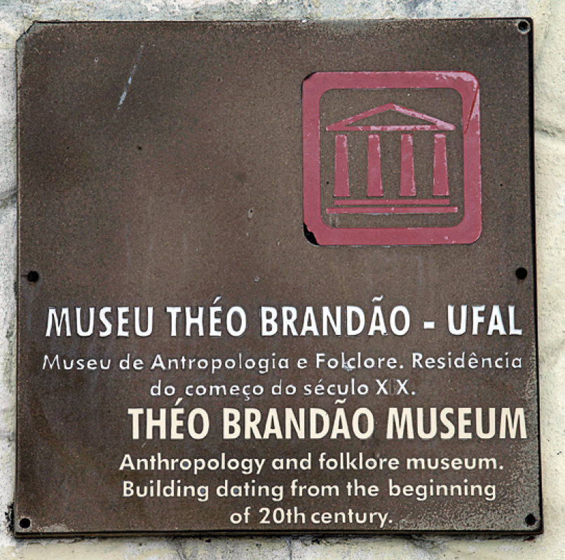 Fotografia. Parede em cinza, com placa em marrom. Na ponta da direita da placa, há um ícone em rosa de um prédio antigo com telhado triangular. Na parte inferior, texto em branco: Museu Théo Brandão, UFAL. Museu de Antropologia e Folclore. Residência do começo do século vinte Brandão Museum, Antropology and folklore museum. Building dating from the beginning of twentieth century.