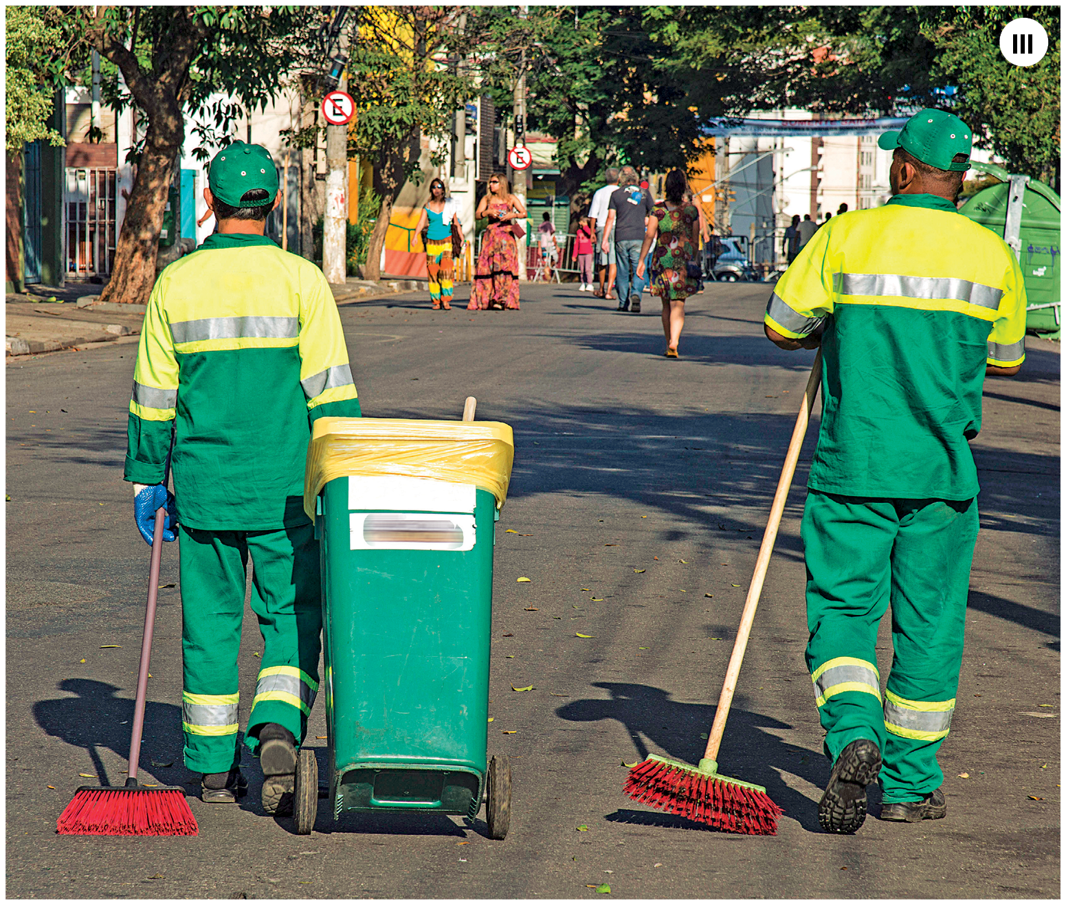 Fotografia três. Dois homens de costas em uma rua. Eles são varredores de rua e vestem uniformes compostos por camisas, calças e bonés em amarelo e verde com faixas reflexivas em cinza no peito, nos braços e na parte inferior das pernas. Eles também usam botas pretas e arrastam vassouras com cerdas vermelhas nas mãos esquerdas. O homem no canto esquerdo usa luvas azuis e com a mão direita puxa uma lixeira em verde com sacola plástica amarela em seu interior. Ao fundo, em segundo plano, pedestres andando, árvores e postes de eletricidade.