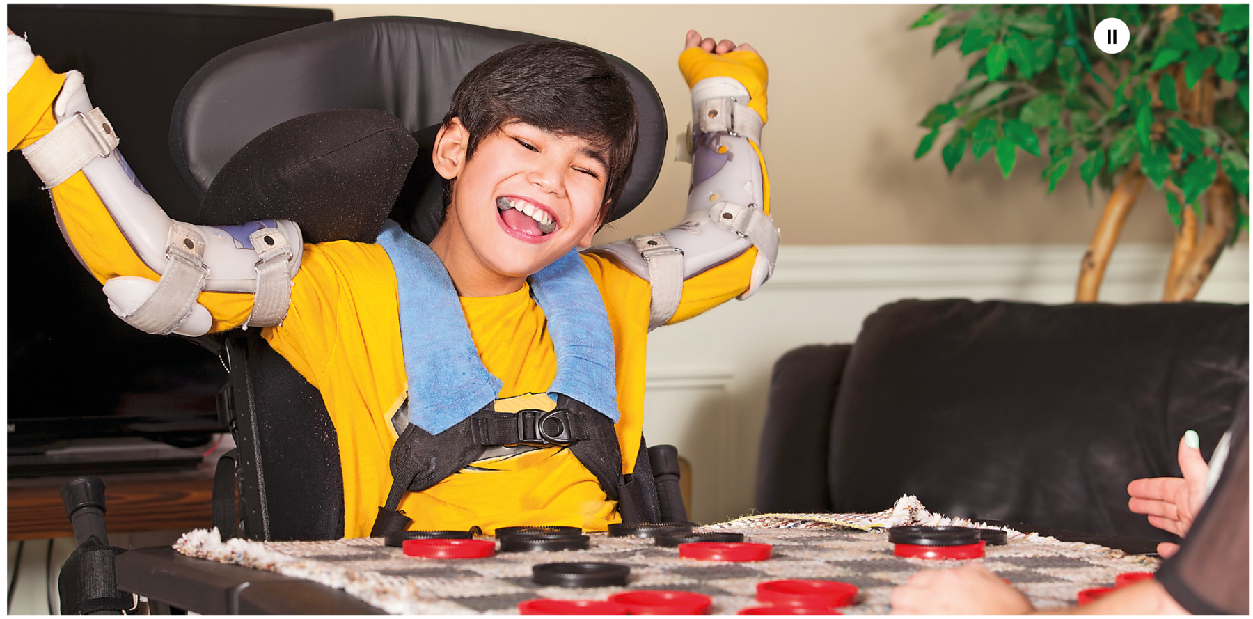 Fotografia dois. Um menino visto da cintura para cima atrás de uma mesa, à esquerda, de frente para uma mesa vestindo camiseta amarela de mangas compridas em uma sala de estar de frente para outra pessoa que está quase totalmente fora de quadro. Ele está sentado em uma cadeira preta, preso com cintos de segurança no tórax e nos braços há próteses de plástico. Ele olha para frente rindo. Sobre a mesa, botões pequenos em preto e vermelho em um tabuleiro de jogo de damas.