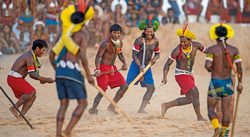 Fotografia. Local com solo arenoso claro, com seis homens indígenas jogando. Eles têm cabelos pretos, sem camiseta e bermuda em azul e vermelho, alguns com cocares sobre a cabeça com penas em amarelo. Todos seguram hastes de madeira que usam para empurrar algo que não está identificado na foto e ao fundo, outras pessoas vistas parcialmente acompanham a partida.