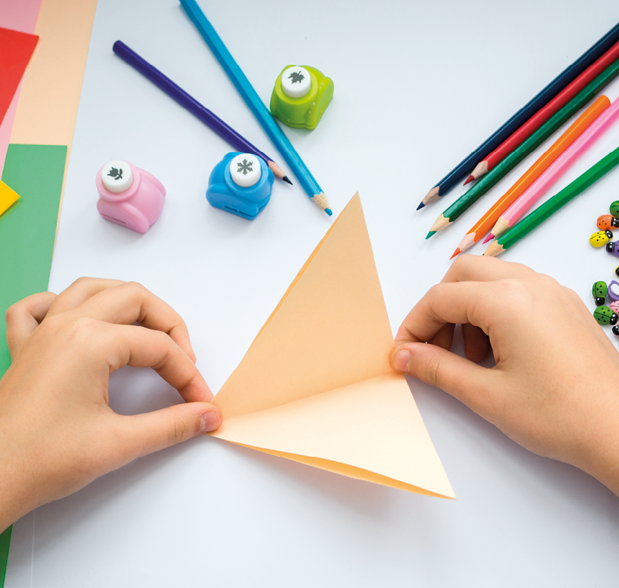 Fotografia. Vista do alto das mãos de uma criança segurando um papel retangular dobrado no meio formando um triângulo, segurando nas pontas e em cima da mesa há lápis de diversas cores e três cortadores de papel pequenos.