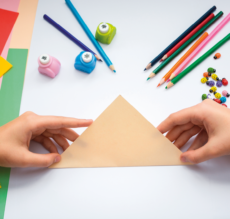 Fotografia. Vista do alto das mãos de uma criança sobre uma mesa, segurando triângulo feito com papel bege dobrado ao meio. Ao centro da folha, uma dobra demarcada. Em cima da mesa há lápis de diversas cores e três cortadores de papel pequenos e folhas de papel em cores diversas.