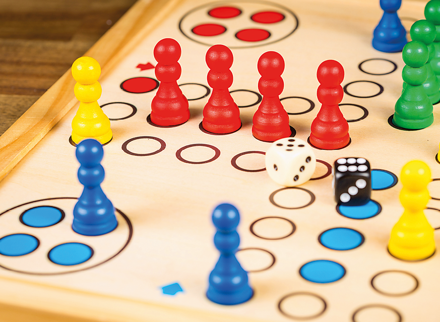 Fotografia. Vista geral de tabuleiro de madeira, com círculos pequenos em linha, em diversas direções, peças de peões coloridos em amarelo, azul, vermelho e verde, com dois dados, um branco e outro preto.