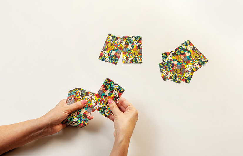 Fotografia. Mãos de uma pessoa segurando várias cartas de costas em tons coloridos de preto, amarelo, vermelho e branco. Perto dela, à direita, dois montes de cartas separados.