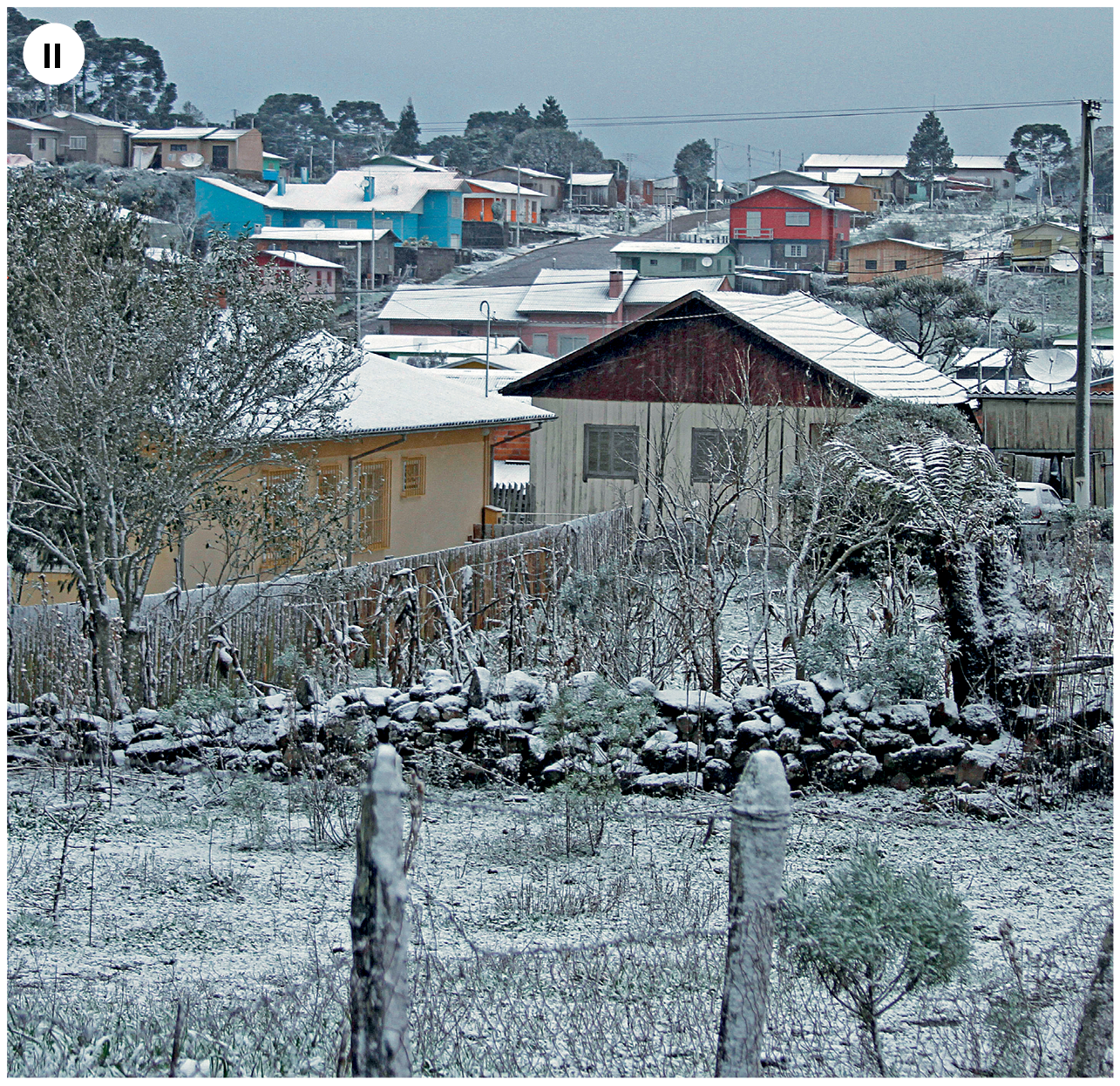 Fotografia dois. Vista geral de terreno com pedras e gramas, com árvore à esquerda, cobertos por fina camada de neve. Em segundo plano, casas e prédios com paredes em bege, azul, vermelho e outras cores e com telhado triangular e uma fina camada de neve em cima. No alto, céu nublado.