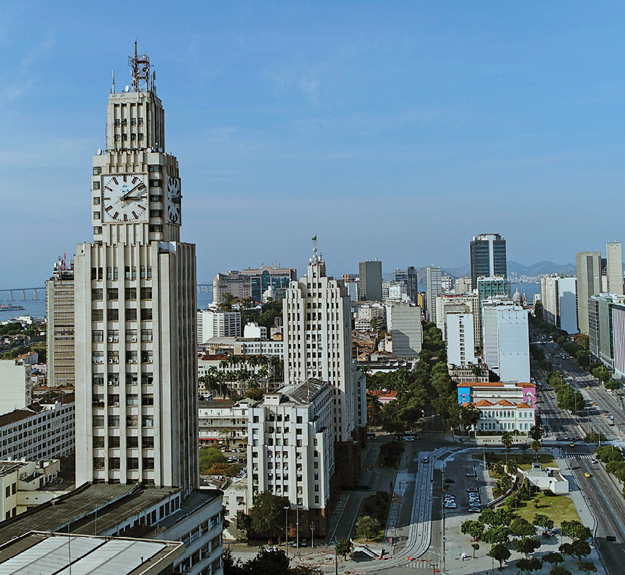 Fotografia. Vista geral de cidade com prédios de diferentes alturas. À esquerda, em primeiro plano, um prédio grande, com um relógio analógico grande no centro e muitas janelas. Entre os prédios, ruas, árvores e mais ao fundo, outras construções. No alto, céu azul sem nuvens.