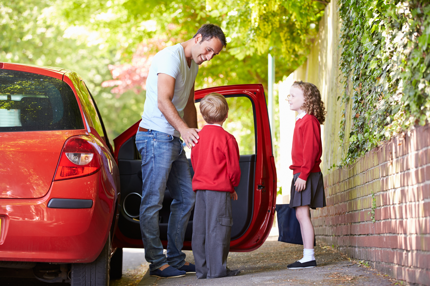 Fotografia. À esquerda, um carro vermelho visto parcialmente, com a porta direita aberta, com um homem em pé, perto de um menino e uma menina. O homem tem cabelos escuros, de camiseta em verde-claro e calça jeans e sorri para o menino. O menino é loiro, de blusa de mangas compridas em vermelho, calça cinza. À direita, a menina está com blusa em vermelho, saia cinza, meias em branco e sapatos pretos. Em segundo plano, muro e árvore e folhas verdes.