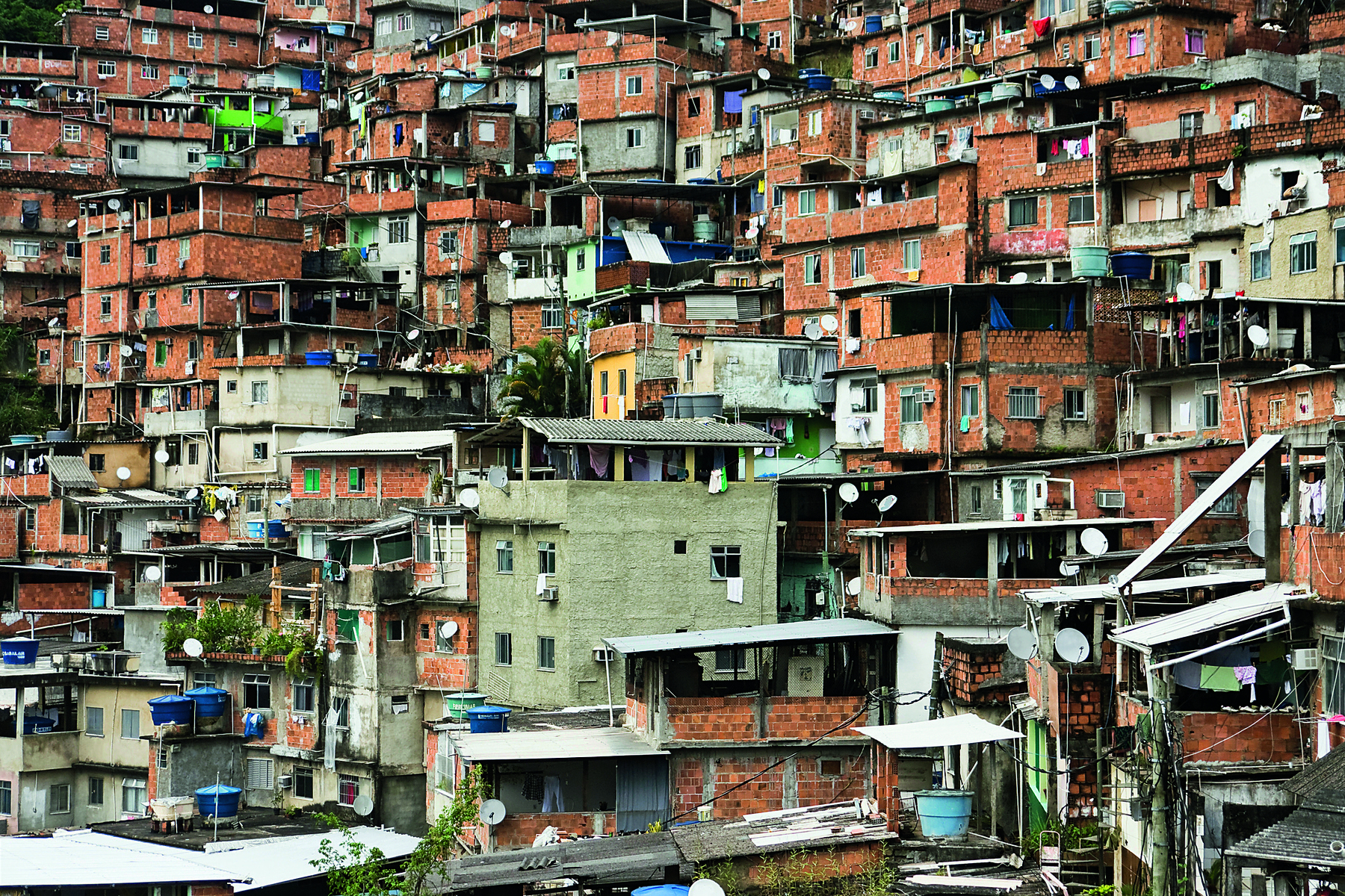 Fotografia. Vista geral de dezenas de casas aglomeradas, em um morro, com paredes em tons de marrom e algumas em cinza, construídas irregularmente.