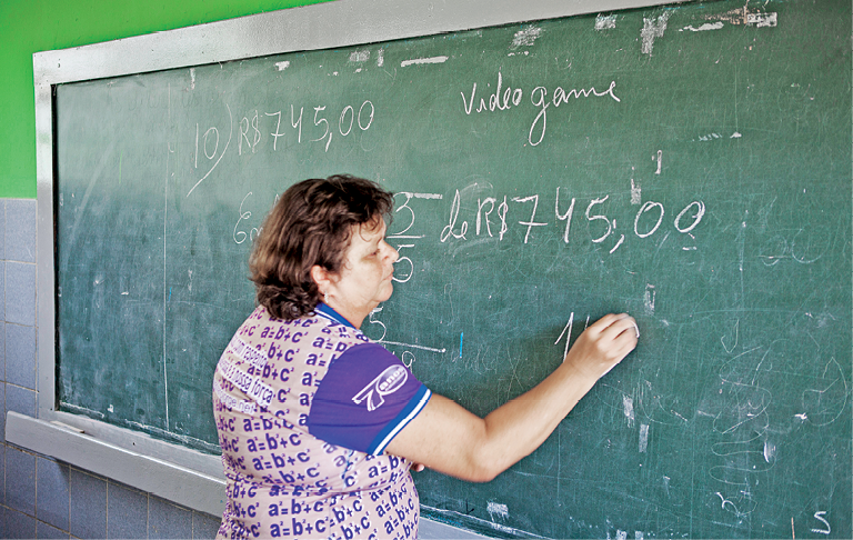 Fotografia. Uma mulher vista de costas, da cintura para cima, de cabelos curtos em castanho, com camiseta em branco e roxo, escrevendo um cálculo em uma lousa com giz amarelo.