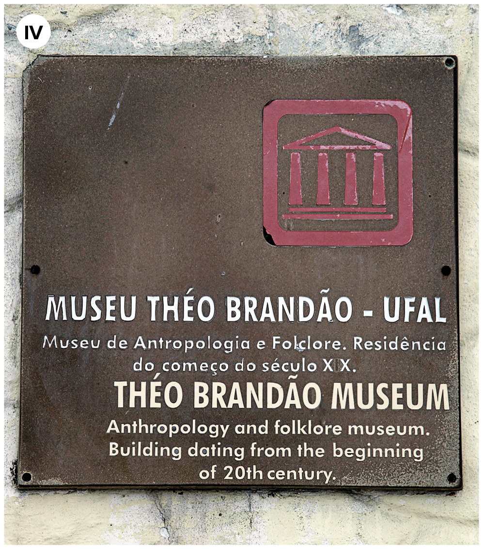 Fotografia quatro. Parede em cinza, com placa em marrom. Na ponta da direita da placa, há um ícone em rosa de um prédio antigo com telhado triangular. Na parte inferior, texto em branco: Museu Théo Brandão, UFAL. Museu de Antropologia e Folclore. Residência do começo do século vinte Brandão Museum, Antropology and folklore museum. Building dating from the beginning of twentieth century.