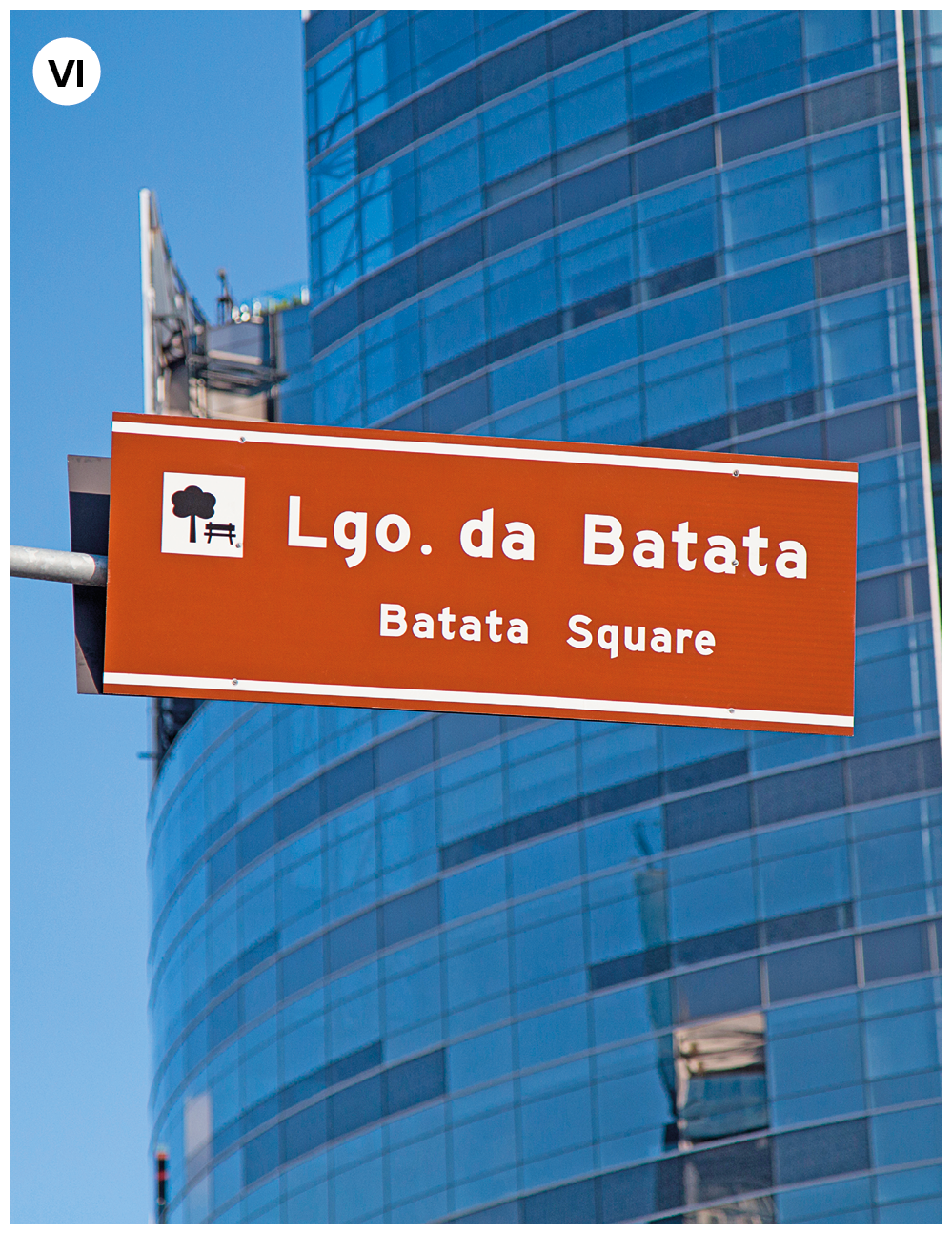 Fotografia seis. Foco na parte superior, placa em marrom com texto em branco: Largo. Da Batata, Batata Square. Ao fundo, vista parcial de um prédio alto espelhado, em tons de azul.