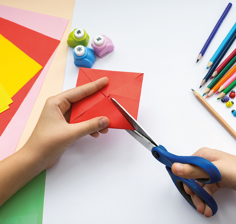 Fotografia. Vista do alto das mãos de uma criança cortando uma dobradura em papel vermelho em formato de quadrado com uma tesoura  e em cima da mesa há lápis de diversas cores e três cortadores de papel pequenos, além de algumas folhas de cores diversas.
