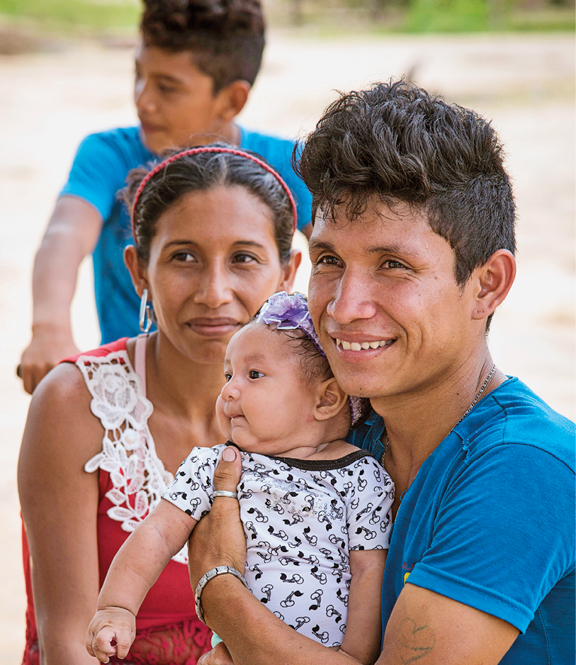 Fotografia. À direita, um homem de cabelos pretos, de camiseta azul, segurando no colo, uma bebê. Ele olha para frente, sorrindo e a bebê, de cabelos pretos, tem uma faixa sobre a cabeça em azul-claro e blusa de mangas curtas em branco e preto. Atrás deles, uma mulher vista da cintura para cima, de cabelos pretos com tiara vermelha sobre a cabeça, blusa regata em vermelho e gola em branco, bordada. Ela olha ao longe, com leve sorriso para a esquerda. Ao fundo, vista parcial de um menino moreno, cabelos escuros, de camiseta azul-claro, olhando para a esquerda.