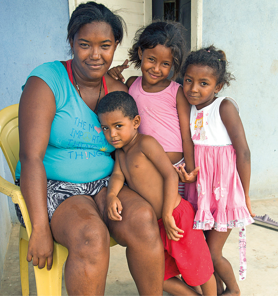 Fotografia. Uma mulher sentada em cadeira de plástico amarela e ao lado dela, três crianças à direita, em pé. A mulher é negra, tem cabelos pretos presos, de blusa de mangas curtas em azul, bermuda em branco e preto, olhando para frente, sorrindo. Perto dela, menino moreno, de cabelos pretos, sem camiseta e com bermuda em vermelho. Na ponta da direita, duas meninas negras de cabelos encaracolados, sendo que uma delas veste regata rosa e a outra uma regata branca e saiote rosa. Ambas olham para frente sorrindo.