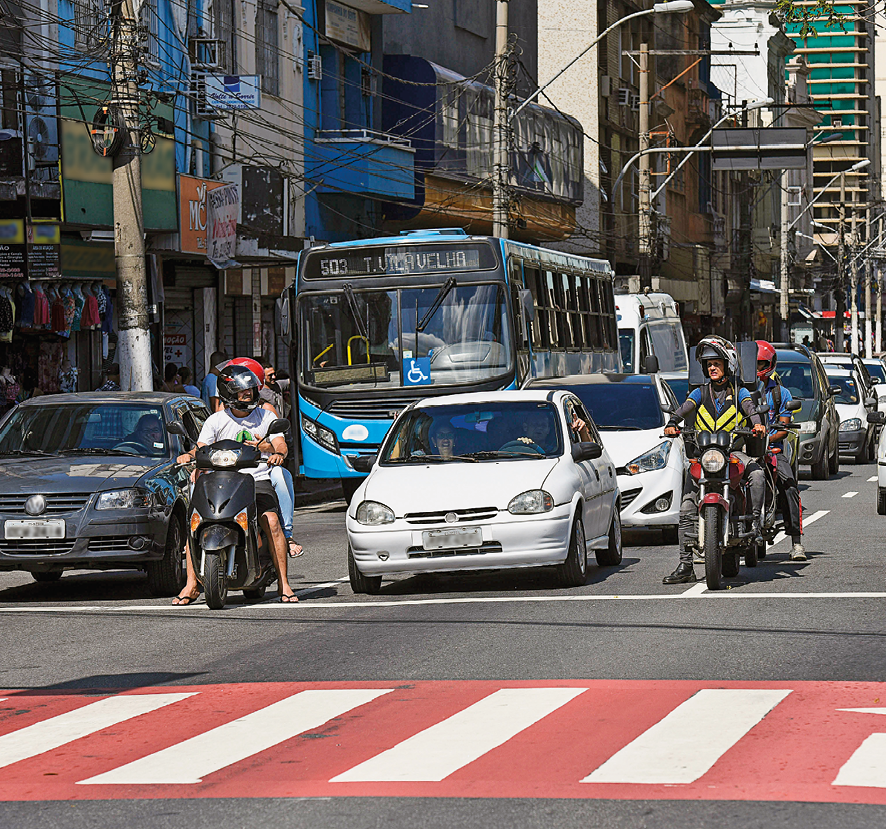 Fotografia. Rua de cor cinza, com faixa de pedestre em vermelho e branco. Ao fundo, três fileiras com automóveis: carros, motos e ao fundo, ônibus azul claro. À esquerda, prédios, comércios e postes de energia.