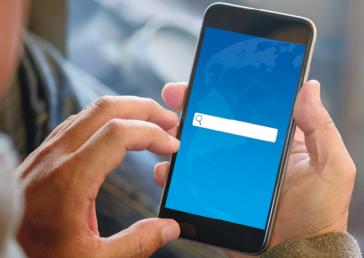Fotografia um. Mãos de uma pessoa segurando na mão direita, um celular preto, com imagem em azul e barra de busca branca sem nada escrito.