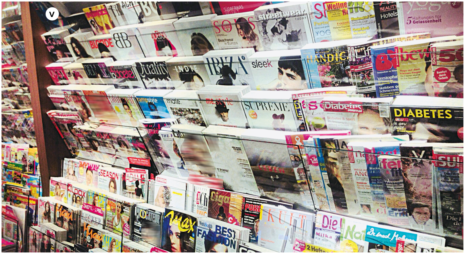 Fotografia cinco. Vista geral de uma parede de madeira de uma livraria ou banca de jornal com oito andares e várias revistas uma ao lado da outra, com fio preto passando pela frente delas e segurando-as no lugar. Há revistas de diferentes títulos, capas e fotos variadas.