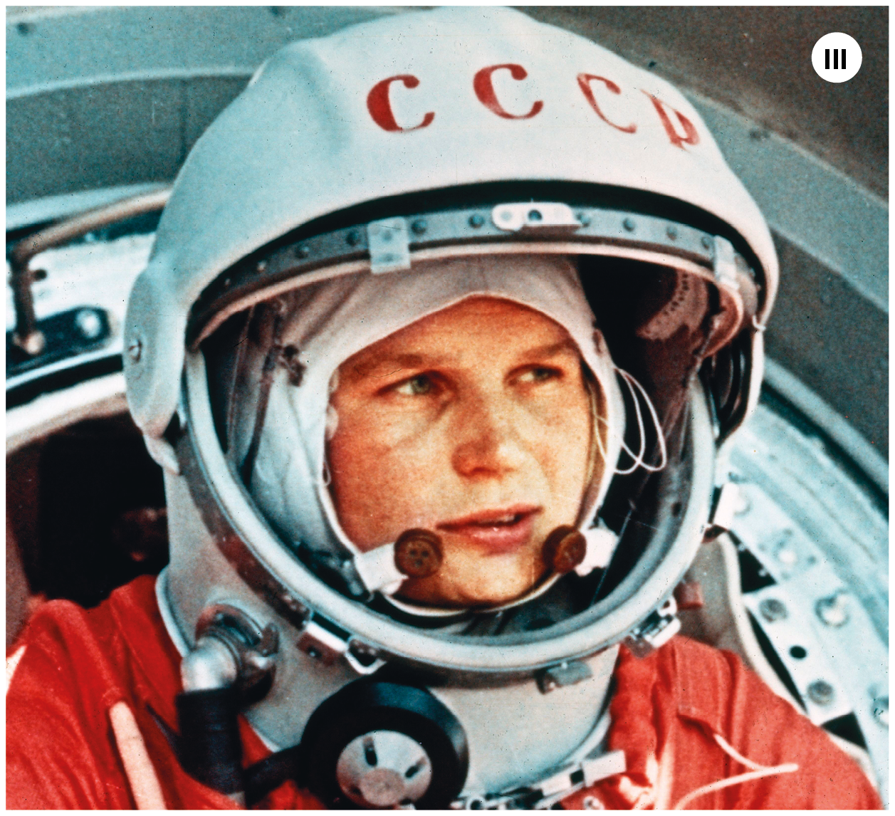 Imagem três. Fotografia. Uma mulher branca vista dos ombros para cima, de cabelos claros, capacete branco e roupa de astronauta com detalhes em branco e vermelho. Ela olha para a direita, ao longe. Ao fundo, objeto arredondado em ferro de cor cinza.