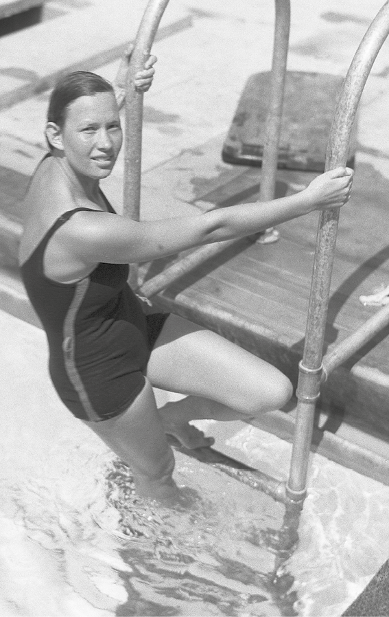 Fotografia. Em preto e branco, vista do alto, de uma mulher branca subindo em escada de piscina. Ela tem cabelos escuros e maiô escuro. Ela olha para frente, segurando os canos laterais da escada.