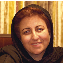 Fotografia. Shirin Ebadi. Uma mulher vista dos ombros para cima, de cabelos longos castanhos, nariz e lábios finos e roupa marrom. Ao fundo, parede amarela. A mulher olha para frente sorrindo.