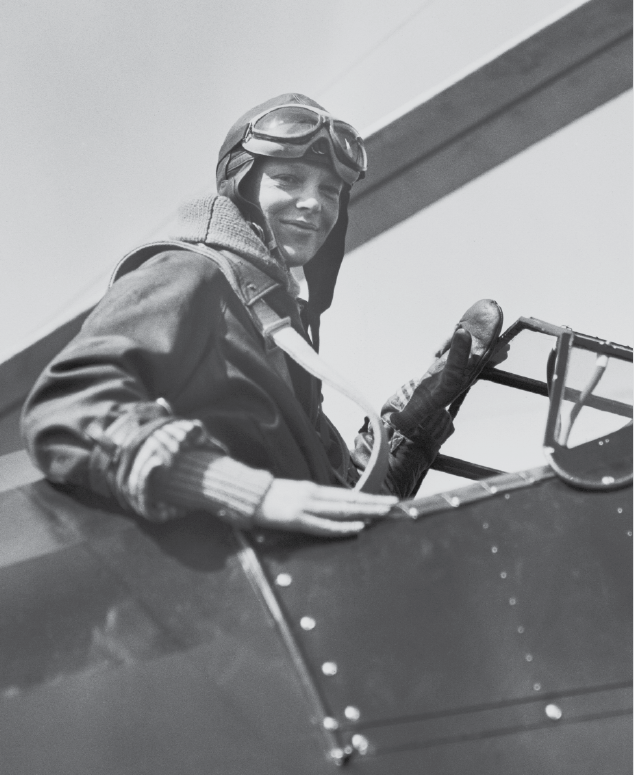 Fotografia. Amelia Earhart em preto e branco. Uma mulher dentro de um avião escuro, antigo. Ela tem chapéu de aviador sobre a cabeça com óculos grandes e laterais longas que cobrem suas orelhas, e blusa de mangas compridas. Ela olha para o lado direito com um leve sorriso.