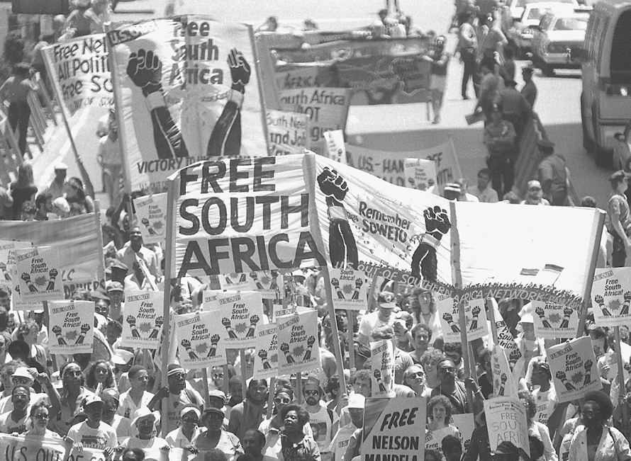 Fotografia. Em preto e branco, vista do alto de rua com dezenas de pessoas negras marchando na rua ao lado de carros. Entre elas, cartazes em branco com textos como: FREE SOUTH AFRICA, REMEMBER SOWETO e FREE NELSON MANDELA.